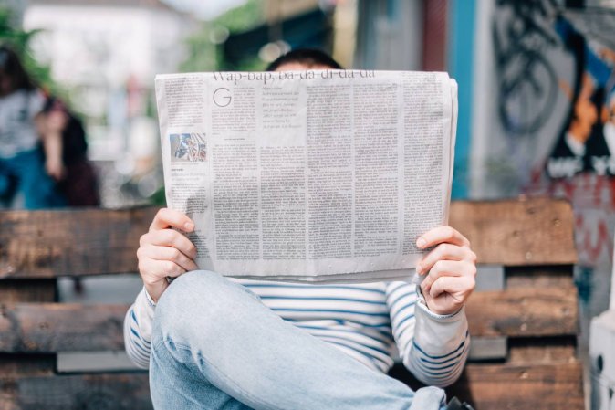 A person reading a newspaper while sitting on a bench outside.
